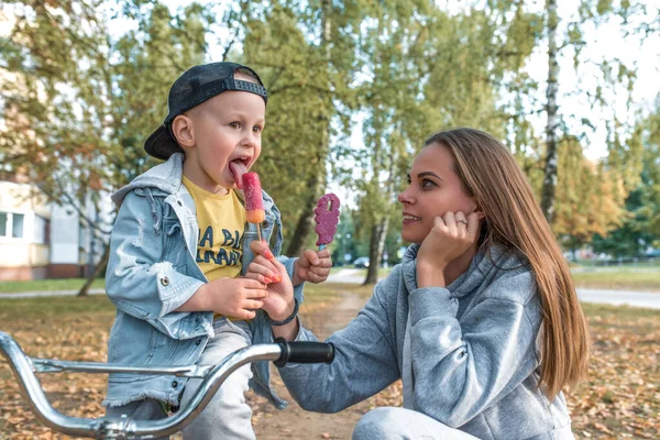 Mom woman little boy son of 4-5 years old, eat ice cream, support care and education, have snack nature, relax in summer park city park. Casual wear denim cap, sneaker pants. Mothers day. — Stockfoto