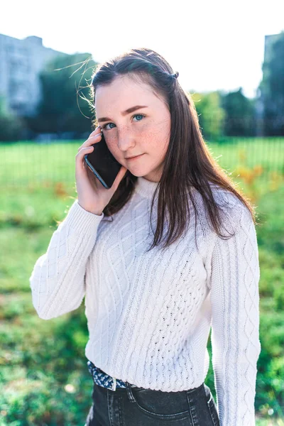 青春期的女学生，夏天穿着白色毛衣站在城市里，打了个电话，背景绿草，在社交网络和互联网上收听应用程序中的语音留言. — 图库照片