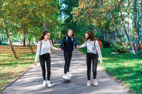 Meninas adolescentes alunas montam um skate depois da escola, sorrindo feliz, emoções de alegria, prazer e diversão, camisolas quentes no outono na rua na cidade. Melhores amigos namoradas . — Fotografia de Stock