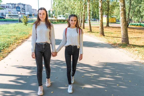 Dua gadis sekolah teman-teman berjalan di kota di musim panas setelah pulang sekolah. Di balik ransel, kasual memakai sweater jeans. Musim gugur, pohon-pohon latar belakang meninggalkan jalan . — Stok Foto