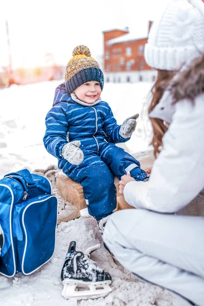 Ibu membantu wanita, musim dingin, memakai sepatu skate, anak laki-laki berusia 3-5 tahun, kereta belajar skate, pakaian hangat, rileks pada akhir pekan. Background snow drifts ice rink. Happy tersenyum ceria dan gembira . — Stok Foto