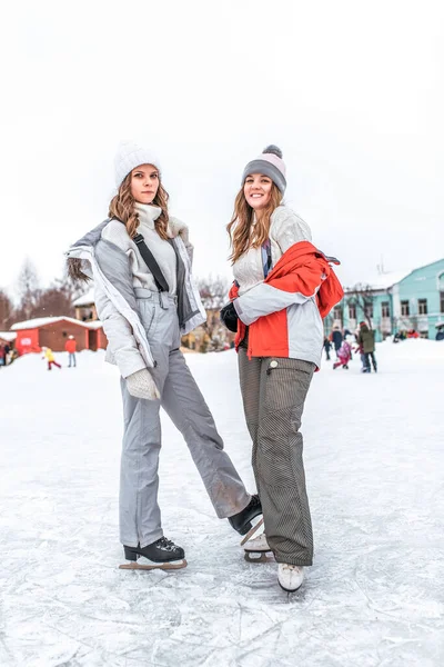 Városi jégpálya, hósodródik hóháttér. Az érzelmek örömet okoznak a boldogságnak. Két lány barátnő téli lovaglás korcsolyapálya sportruházat meleg ruhák, pulóver kezeslábas kalap. Boldog mosolygást, pihenő hétvégét.. — Stock Fotó