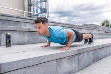 Erkek sporcu yaz aylarında öne doğru itiyor. Arka planda ise basamaklar var. Aktif gençlik yaşam tarzı, doğada spor. Cesaret ve motivasyon gücü. Su ve proteinli titretici..