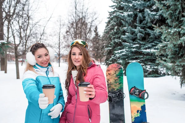 Due ragazze amiche inverno parco resort tenere fuori tazze di tè caldo caffè, offrire e trattare loro condividere. Emozioni piacere comfort. Tute sportive, sfondo alberi di neve snowboard. Spazio libero per il testo . — Foto Stock