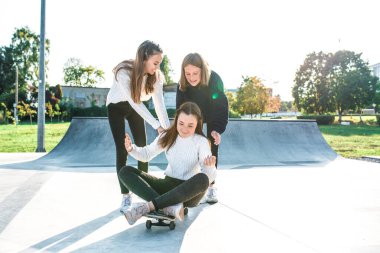 Üç genç kız kaykaya biniyor, mutlu mesut oynuyor ve gülüyorlar, yazın spor sahasında, sonbahar öğleden sonra sıcak kazaklarla. Metni kopyalamak için boş alan.