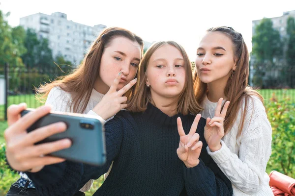 Tres chicas adolescentes. Se toman fotos en el teléfono, sonrisas felices juegan y tontean. Aplicación en línea en Internet, redes sociales. En la ciudad de verano, suéteres cálidos otoño . —  Fotos de Stock