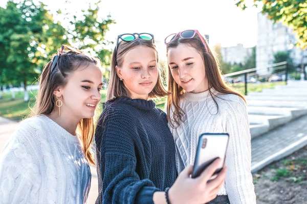 Три девочки-подростки, подростки в летнем городе отдыхают после школы, в руках смартфона, читают сообщения и пишут видео-звонок, онлайн-приложение в социальных сетях. Счастливы улыбаются . — стоковое фото