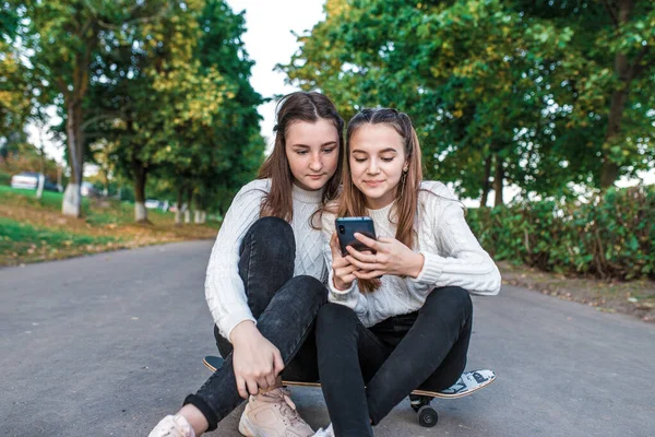 Dos adolescentes adolescentes, mejores novias, parque de verano, monopatín sentado, viendo videos divertidos smartphone, sonrisa feliz, ropa casual, día de otoño. Relajación conceptual después de la universidad escolar . —  Fotos de Stock