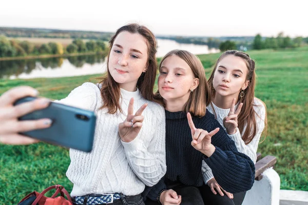 Tři dospívající dívky, teenageři v letním městě mají odpočinek po škole, ruce smartphone, selfie video hovor on-line aplikace na sociálních sítích. Šťastný úsměv. Nenucené oblečení, svetr. — Stock fotografie