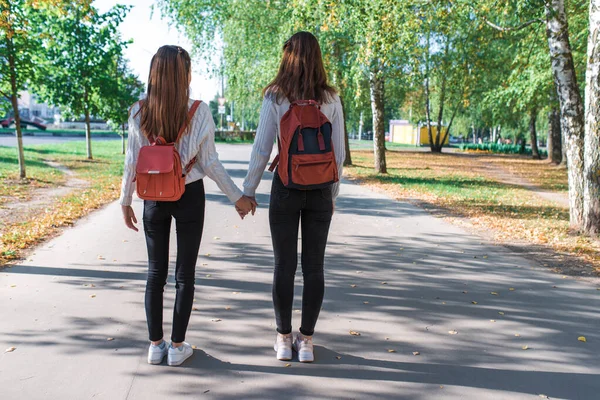 Dua gadis remaja berjalan di musim panas di taman, berjalan sepulang sekolah dan kuliah, bergandengan tangan, sahabat pulang ke rumah setelah berlatih dengan ransel. Ruang kosong untuk menyalin teks . — Stok Foto