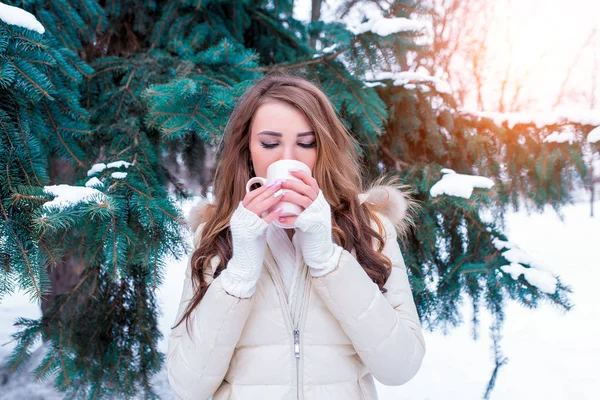 Chica en invierno bebe una bebida caliente de una taza, fin de semana en el complejo, árboles verdes de fondo ventisqueros. Con una chaqueta caliente, puños y una bufanda. Té de café caliente en clima frío . — Foto de Stock