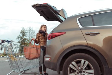 Yaz mevsiminde kadın otoparkta, bagajı açık, alışveriş merkezini çantalarla dolduruyor, alışveriş arabalarını dükkandan alıyor, kapalı devre otomatları kullanıyor. Deri ceket ve çanta..