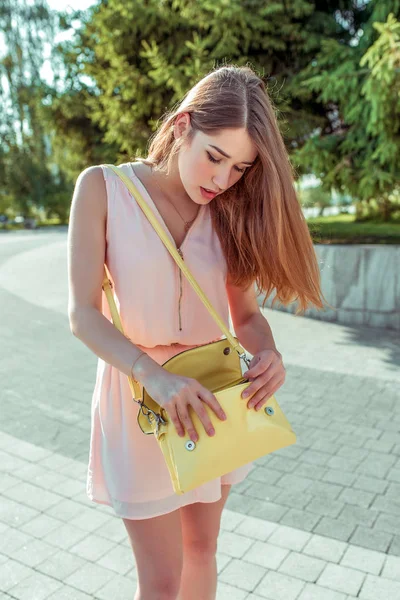 Niña en vestido rosa bolso amarillo, en la ciudad soleada día de verano, en busca de llaves y teléfono inteligente, olvidó y perdió cosas en casa. Sorpresa y frustración. Figura bronceada pelo largo . — Foto de Stock