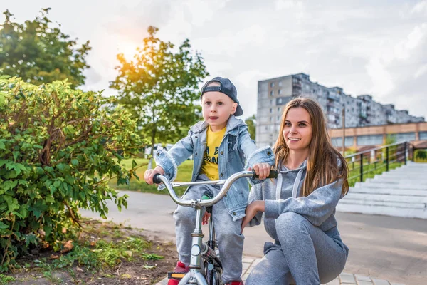 年轻母亲教女人骑自行车，与3-5岁的小男孩玩耍，夏天在城市公园里，秋天穿上大自然的衣服，感受柔情和呵护. — 图库照片