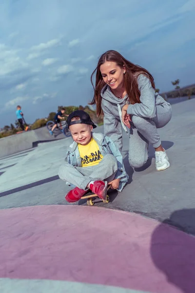 Ung mor med liten pojke son, lycklig familj lär sig att rida skateboard, sommar utomhus, lekplats. Känslor av glädje, glädje och glädje. Vårdstöd vid utbildning, föräldraskap. — Stockfoto