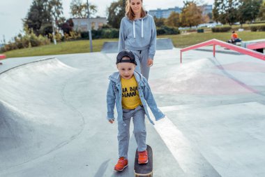 Küçük oğlu olan genç bir anne, mutlu bir aile kaykay sürmeyi öğreniyor, yazın açık havada, sonbahar gününde. Zevkin duyguları, oyun parkının eğlencesi. Eğitimde bakım desteği, ebeveynlik.