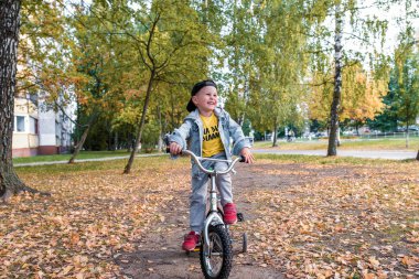 3-5 yaşındaki küçük mutlu çocuk, yazın şehir parkında, gülümser, sevinir, bisiklet sürmek için çok çalışır. Sonbahar giysileri. Zevk ve rahatlama duyguları. Gülün ve eğlenin..