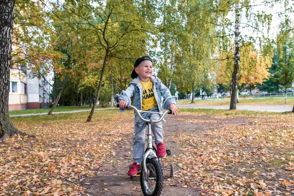 Anak kecil yang bahagia berusia 3-5 tahun, di musim panas di taman kota, tersenyum, bersukacita, bekerja keras untuk mengendarai sepeda. Pakaian musim gugur. Emosi kesenangan dan relaksasi. Tertawa dan bersenang-senang . — Stok Foto
