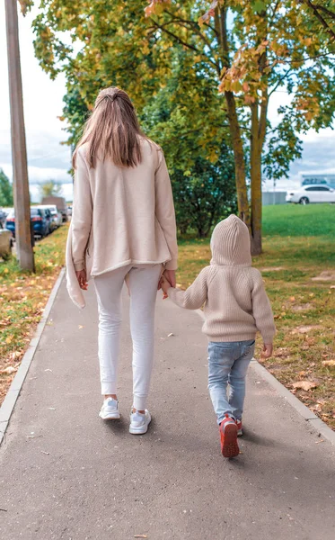 Kadın anne sokakta yürüyor, küçük oğluyla el ele tutuşuyor, sonbaharda şehirde yürüyor, sokak boyunca yürüyor, arka plan ağaçları, sıcak günlük kıyafetler, kapüşonlu bej kazak.. — Stok fotoğraf