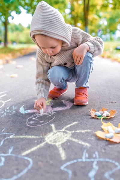 Sonbaharda sokakta küçük bir çocuk, kaldırıma resim çiziyor, renkli pastel boyalar. Soğuk bir günde sıcak kıyafetler, kapüşonlu bej bir hırka. Hafta sonu molaları, yaratıcılık ve çizimler.