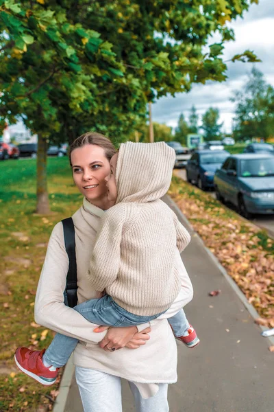 Kadın anne sokakta yürür, küçük oğlunu kucağına alır, sonbahar şehri, cadde boyunca yürür, arka plan ağaçları, sıcak günlük kıyafetler, kapüşonlu bej kazak. Soğuk hava esintidir.. — Stok fotoğraf