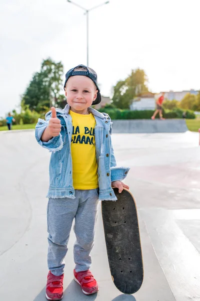 Glad liten pojke 3-5 år gammal, på sommaren på idrottsplats i staden, tummen upp, leenden, glädje, avskedsord undervisning på skateboard. Höstkläder. Känslor av nöje och avkoppling. — Stockfoto