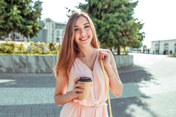 Щаслива дівчина сміється з посмішок, стоїть у місті влітку, тримає чашковий кавовий чай своїми руками. Рожеве плаття довге волосся повсякденний макіяж, фон з дерев. Емоції радості релаксації вихідні . — стокове фото
