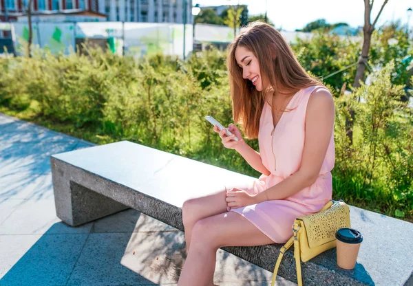 Gaun merah muda yang cantik, taman kota musim panas duduk di bangku cadangan, smartphone di tangannya, membaca pesan kepada Internet, aplikasi online di jaringan sosial. Happy tersenyum, ruang kosong untuk teks . — Stok Foto