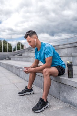 Erkek sporcu oturur, cep telefonuyla el ele tutuşur, müzik kulaklığı dinler, yaz antrenmanından sonra şehirde dinlenir, arka plan bulutu adımları. Aktif gençlik yaşam tarzı, zindelik doğası.