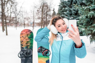 Güzel bir kadın, cep telefonuyla konuşan video konuşması, resim selfie 'si. Spor tulumu. Snowboard arka planda kar, Noel ağaçları. Duygular mutluluk gülümseme kış tatili. Boş alan metni.