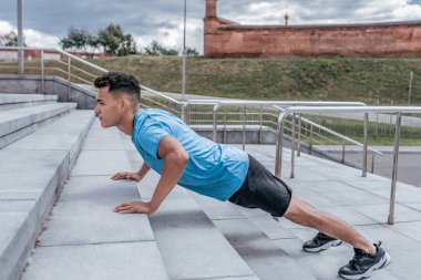 Atletik adam, beton basamaklardan şınav, şehirde yaz öğleden sonra eğitimi, aktif yaşam tarzı, modern fitness antrenmanı, spor tişörtü şortu spor ayakkabıları..