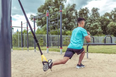 Atletik adam kemer askısı ile egzersiz yapar, bacak kaslarını çalıştırır, yazın şehirde egzersiz yapar, aktif yaşam tarzı, modern fitness egzersizi. Motivasyon metni için boş alan.