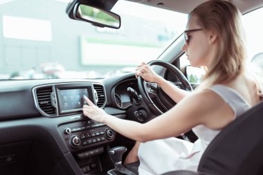 Kadın araba kullanıyor, dokunmatik ekranda parmağa basıyor, internetten uygulama seçiyor, radyo navigasyonunu etkinleştiriyor ve şehirdeki güzergahı arıyor. Arkaplan otoparkı.
