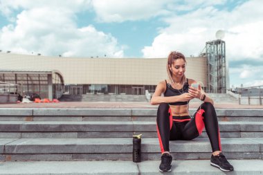 Kız sporcu, yaz günü spor salonunda merdivenlerde oturur. İnternette mesajlar yazıyor, akıllı telefon müzik kulaklıkları dinliyor. Podcast konferansını dinlemekten hoşlanıyor. Aktif yaşam tarzı uygunluğu, şık kadın..