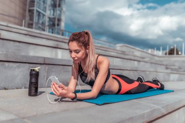 Kız sporcu yoga jimnastiğine yatar, telefona bakar, mesaj yazar, müzik kulaklığı dinler, yaz günü İnternet 'te dinlenir. Podcast konferansını dinlemekten hoşlanıyor. Etkin yaşam tarzı uygunluğu.