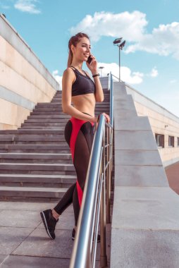 Kız sporcu mutlu bir şekilde telefonda konuşuyor, eğitimden sonra dinleniyor, yaz günü şehirde. Yürümeyi seviyor, internet uygulamasından mesaj dinliyor. Aktif yaşam tarzı uygunluğu, modaya uygun bir kadın..