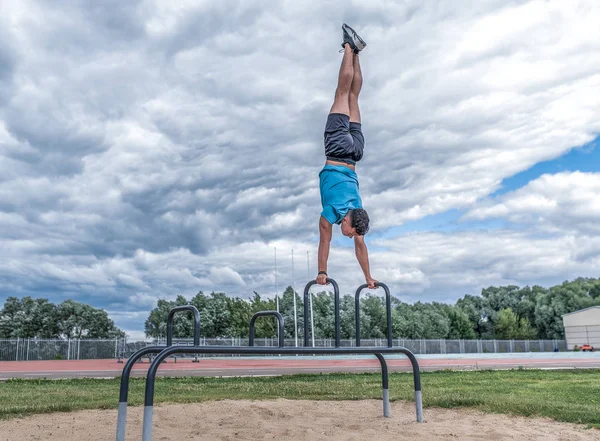 Hombre deportivo de pie de manos, equilibrio, flexiones barra horizontal, ejercicio en la ciudad durante el verano, estilo de vida activo, entrenamiento de fitness moderno. Espacio libre para texto de motivación. Nubes de fondo y campo deportivo . —  Fotos de Stock