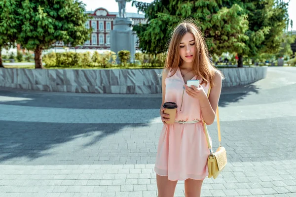 Meisje in roze jurk, staat in de stad in de zomer, kijkt naar de telefoon, leest een bericht online op internet, een kopje koffie thee, social networking applicatie. Vrije ruimte voor tekst. — Stockfoto