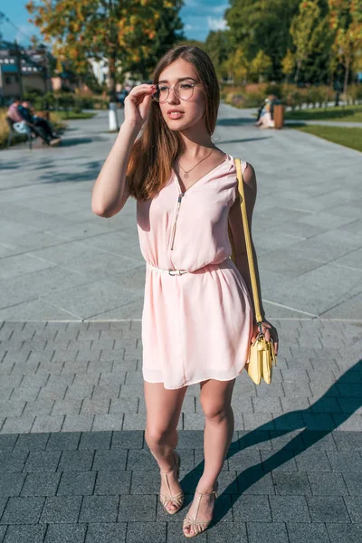 Hermosa muchacha en el vestido rosado, corrige las gafas, en verano en la ciudad, el estilo de vida a la moda y moderno. Bolso sobre el hombro, una figura bronceada . — Foto de Stock