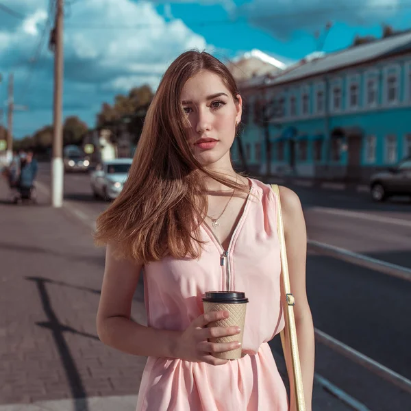 美しい女の子ピンクのドレス、コーヒーティーとカップの手の中の背景の道路の建物、都市で夏には、ファッショナブルな、現代的なファッションライフスタイル、日焼けした姿. — ストック写真