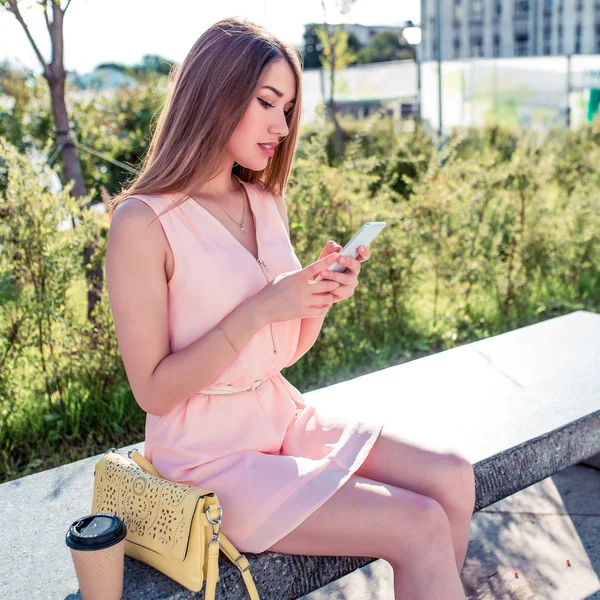 Hermosa niña vestido rosa, se sienta banco del parque, escribe lee el mensaje de teléfono móvil, aplicación en Internet, las redes sociales de la ciudad de verano, el estilo de vida de moda moderna. Copa de bolso con té de café . —  Fotos de Stock