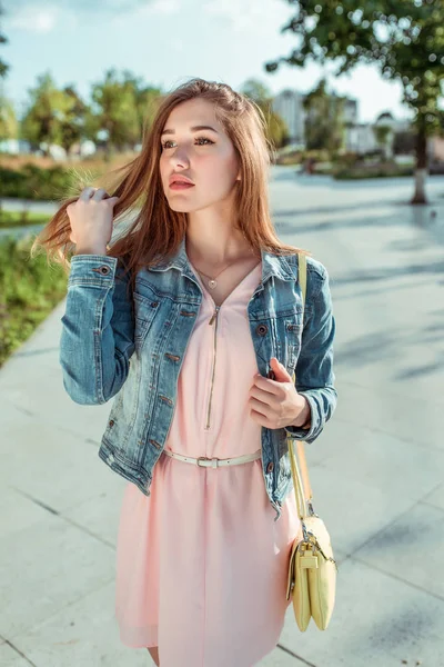 Mooie meisje student in een roze jurk en jeans straalt haar haar, de zomer in de stad, modieuze en moderne mode levensstijl. Gele schoudertas. — Stockfoto