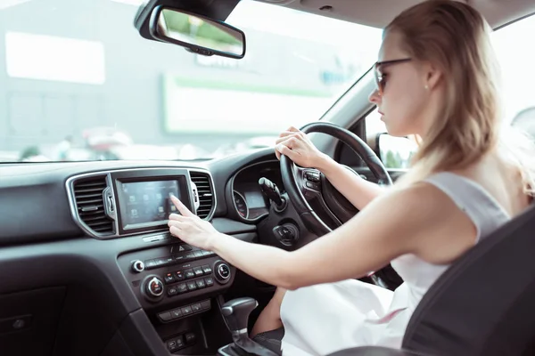 Donna guida auto, preme il dito sul touch screen, seleziona l'applicazione su Internet, attiva la navigazione radio e cerca il percorso in città. Sfondo parcheggio centro commerciale . — Foto Stock