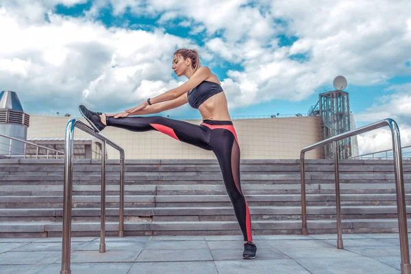 Deri taytlı kız sporcu, antrenmandan önce bacak kaslarını esnetiyor, yaz sabahı şehirde. Spor giysiler, spor ayakkabılar. Aktif yaşam tarzı uygunluğu, modaya uygun bir kadın..