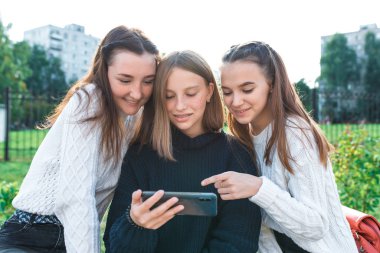 Üç liseli kız öğrenci, 13-15 yaşlarında, sonbahar yaz günü, telefonda videolar izliyor, gülümsüyor, gülüyor, tatilden sonra rahat elbiseler giyiyor. İnternetteki çevrimiçi sosyal ağ uygulaması.
