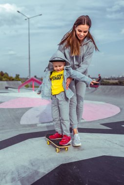 Kadın anne kaykay öğretiyor, destek eğitimi veriyor, küçük oğlan çocuğu, yazın şehirde trenler kaykay öğreniyor, sonbaharda spor sahasında mutlu bir şekilde gülümseyerek..