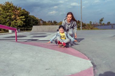 Kadın anne kaykay dersi veriyor, destek eğitimi veriyor, 3-5 yaşlarında küçük bir oğlan çocuğu, yaz şehrini eğitiyor, kaykayda oturuyor, sonbaharda spor sahasında, mutlu bir şekilde gülümsüyor..