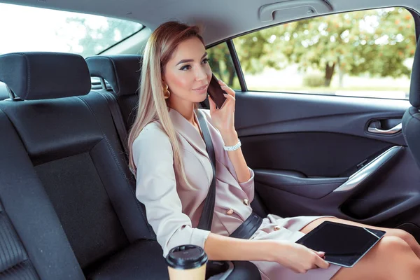 Gyönyörű nő autó belső utas, telefon, dolgozó út, tabletta érintőképernyő. Business Vip taxi kölcsönző autómegosztó város. Nyári lány őszi város. Szigorú öltöny, hosszú haj alkalmi smink.. — Stock Fotó