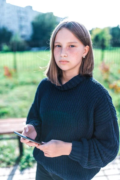10代の女の子14-15歳,秋の日の夏の都市で,スマートフォンを保持,深刻な集中,カジュアル服で,セーター, 10代の女の子の屋外の肖像画. — ストック写真