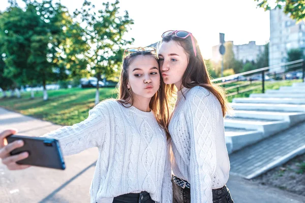 Dos muchachas las colegialas los adolescentes de 12-15 años, en la ciudad otoñal veraniega, en las manos del smartphone, la foto selfie, la videollamada, los jerseys en la ropa de cada día. Emociones alegría, diversión, feliz sonrisa . — Foto de Stock